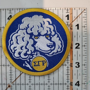 Könnte beinhalten: Ein gestickter Aufnäher in Blau und Gelb mit einem weißen Pudel mit gelben Augen und einem gelben Kreis mit dem Text "ΣΓΡ" in der Mitte.