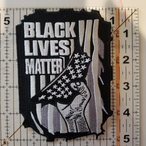 May include: Black Lives Matter patch with a raised fist holding an American flag. The flag is black and white with a gray background. The patch is approximately 2 inches by 3 inches.