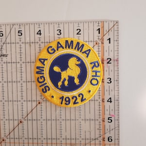 May include: A yellow and blue embroidered patch with the text "Sigma Gamma Rho 1922". The patch features a stylized image of a dog inside a circle.