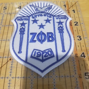May include: A white and blue embroidered patch with a shield shape. The shield has a blue bird with outstretched wings at the top, three blue stars, and the letters "ZOB" in blue. The shield also has two blue torches and the number "1920" in blue.