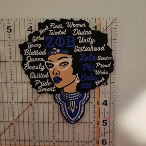 May include: Embroidered patch featuring a black woman with an afro, wearing glasses and a blue and white patterned necklace. The afro is surrounded by text that reads "Black Finer Women", "Talented", "Divine", "Young", "Gifted", "ZΦB", "Unity", "Sisterhood", "Blessed", "Queen", "Beauty", "Skilled", "Pride", "Smart", "Zeta Service", "Phi", "Proud", "Woke", "Beta", "1920", and "Black".