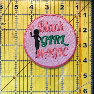 May include: Pink round embroidered patch with a black silhouette of a woman with a flower in her hair. The text "Black Girl Magic" is embroidered in pink and green.