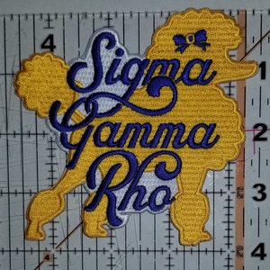 May include: An embroidered patch in the shape of a poodle with a bow on its head. The poodle is yellow and the text "Sigma Gamma Rho" is in purple cursive lettering.