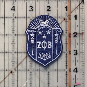 May include: A blue and white embroidered patch with a shield shape. The shield has a white dove with outstretched wings, five white stars, two white torches, and the letters "ZOB" in white. The shield also has the text "1920" in white.