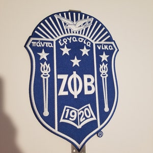 May include: A blue and white embroidered patch with a shield shape. The shield has a white dove flying above it, three white stars, two white torches, the letters "ZΦB", and the number "1920". The text "πάντα ἔργασιά νίκα" is written in white around the top of the shield.