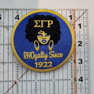 May include: A blue and gold embroidered patch featuring a black silhouette of a woman with an afro, wearing large gold earrings. The text "ΣΓΡ" is above the woman's head and the text "RHOyalty Since 1922" is below her.