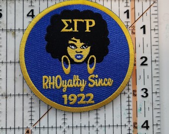 May include: A blue and gold embroidered patch with a black silhouette of a woman with an afro, wearing large gold earrings. The text "ΣΓΡ" is above the woman's head and the text "RHOyalty Since 1922" is below her.