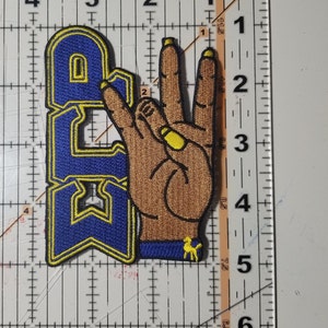 May include: Blue and yellow embroidered patch with the letters "AC" and a brown hand making the "peace sign" gesture.
