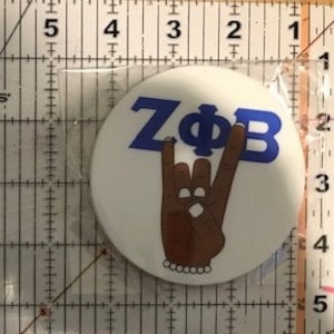 May include: White round button with a blue and white "ZΦB" logo and a brown hand making a rock and roll hand gesture with a pearl bracelet.