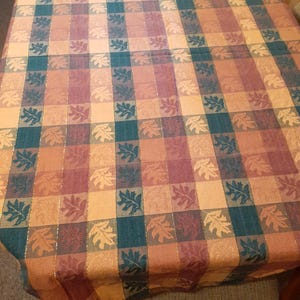 May include: A rectangular tablecloth featuring a plaid pattern in shades of teal, burgundy, and tan. The fabric is decorated with a repeating leaf motif throughout the squares and includes thin, vertical metallic gold threads woven into the design.