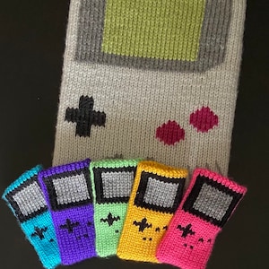 May include: Six knitted Game Boy style keychains in different colors, including blue, purple, green, yellow, pink, and gray. The keychains are hanging from a gray knitted Game Boy background.