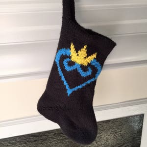 May include: A black knitted Christmas stocking with a blue and yellow heart design and a yellow crown on top.