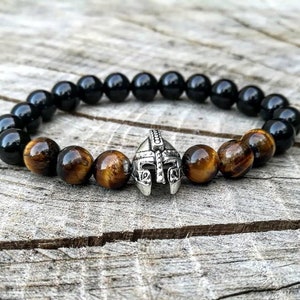 May include: A beaded bracelet featuring black onyx and tiger's eye beads. A silver-colored Spartan helmet charm is centered on the bracelet. The bracelet is displayed on a weathered wooden surface.