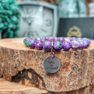 May include: A bracelet featuring spherical beads in shades of purple, green, and white. A silver charm with the words "Why so serious?" is attached. The bracelet is presented on a wooden surface.