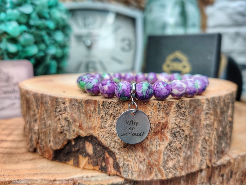 May include: A bracelet featuring spherical beads in shades of purple, green, and white. A silver charm with the words "Why so serious?" is attached. The bracelet is presented on a wooden surface.