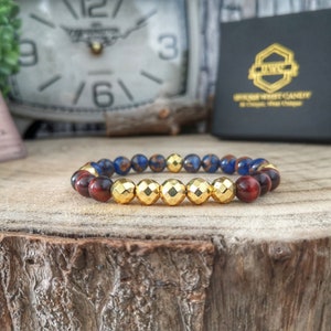May include: A gold, red, and blue beaded bracelet with a gold metal charm. The bracelet is on a wooden surface.