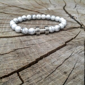 May include: White and gray beaded bracelet with a silver dumbbell charm. The bracelet is on a wooden surface.