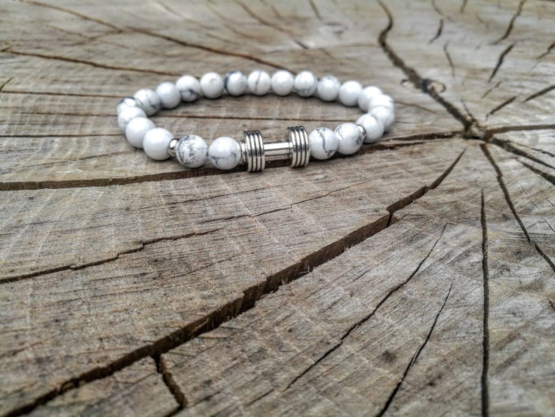 May include: White and gray beaded bracelet with a silver dumbbell charm. The bracelet is on a wooden surface.