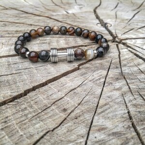 May include: A brown and black beaded bracelet with a silver dumbbell charm. The bracelet is on a wooden surface.
