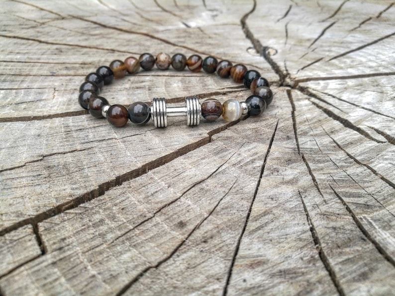 May include: A brown and black beaded bracelet with a silver dumbbell charm. The bracelet is on a wooden surface.