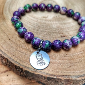 May include: A bracelet with marbled purple, green, and white beads. A silver-tone charm with a Joker illustration is attached. The bracelet is presented on a wooden surface.