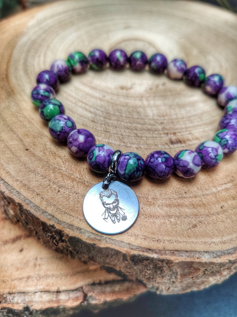 May include: A bracelet with marbled purple, green, and white beads. A silver-tone charm with a Joker illustration is attached. The bracelet is presented on a wooden surface.
