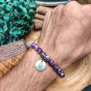 May include: A bracelet featuring speckled purple and green beads. A silver charm with an etched design is attached. The bracelet is worn on a wrist.