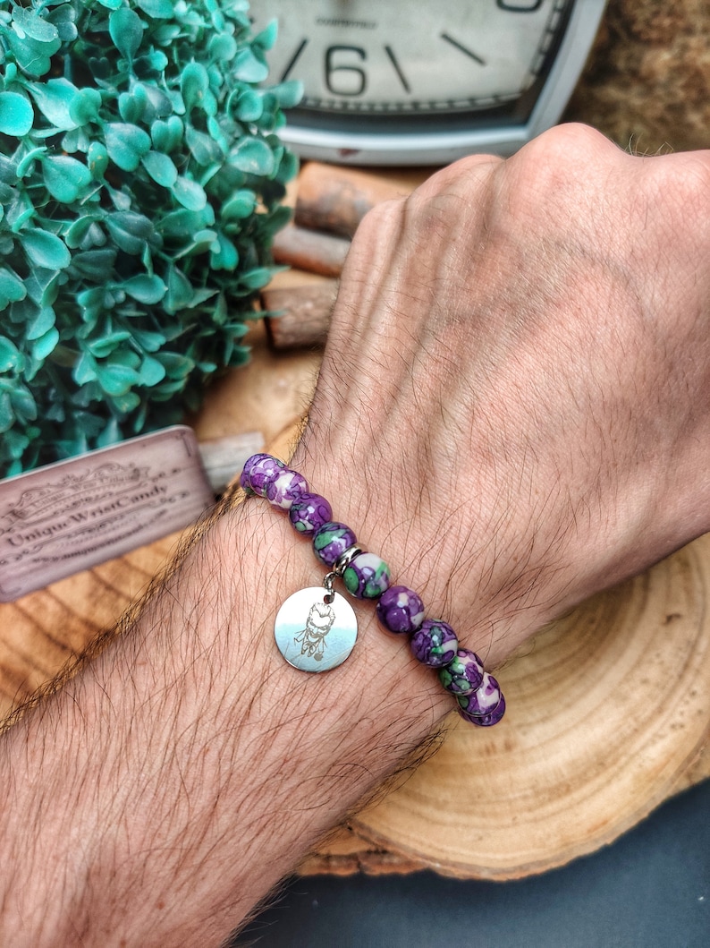 May include: A bracelet featuring speckled purple and green beads. A silver charm with an etched design is attached. The bracelet is worn on a wrist.