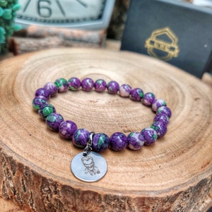 May include: A bracelet with purple and green patterned beads. A silver charm with a character design is attached. The bracelet is presented on a wooden surface. The beads are approximately 1 cm in diameter.