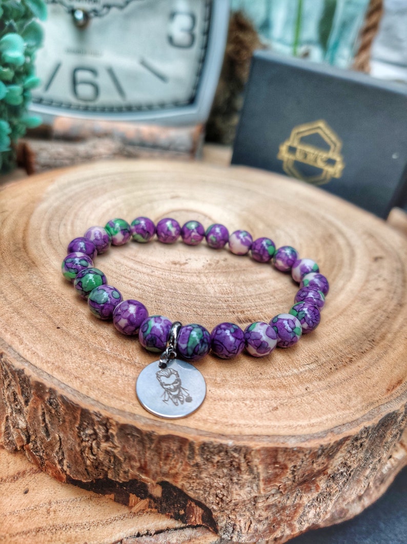 May include: A bracelet with purple and green patterned beads. A silver charm with a character design is attached. The bracelet is presented on a wooden surface. The beads are approximately 1 cm in diameter.