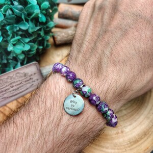 May include: A bracelet featuring purple and green beads and a silver charm. The charm is engraved with the words "Why so serious?". The bracelet is displayed on a wrist.