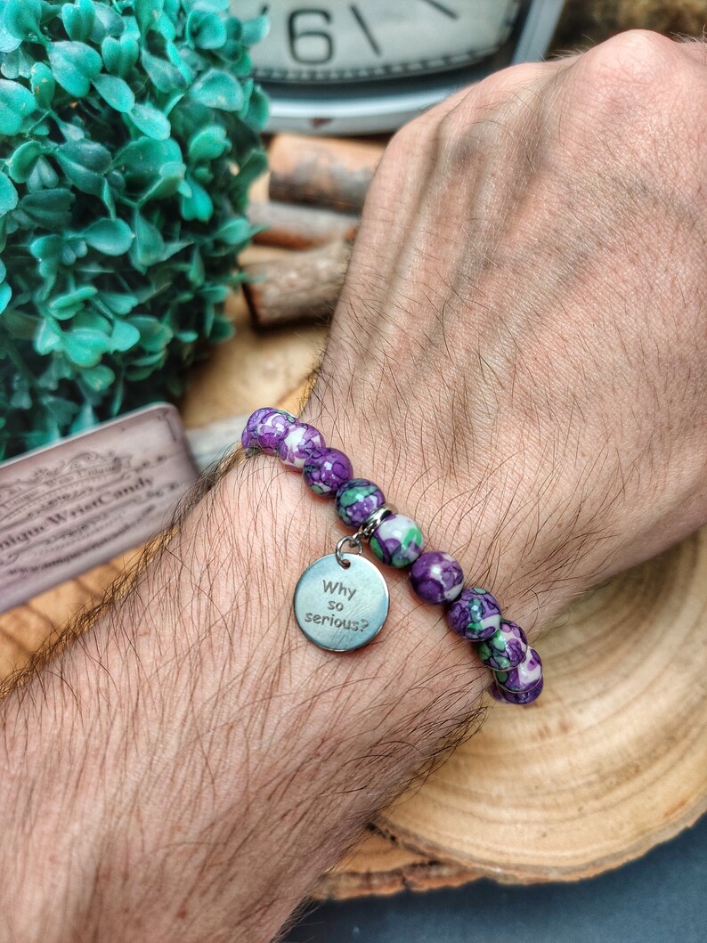 May include: A bracelet featuring purple and green beads and a silver charm. The charm is engraved with the words "Why so serious?". The bracelet is displayed on a wrist.