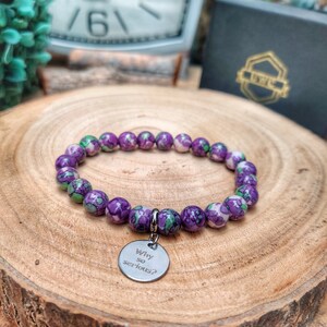 May include: A bracelet with purple and green speckled beads. A silver charm with the words "Why so serious?" is attached. The bracelet is presented on a wooden surface. The beads are approximately 0.8 cm in diameter.