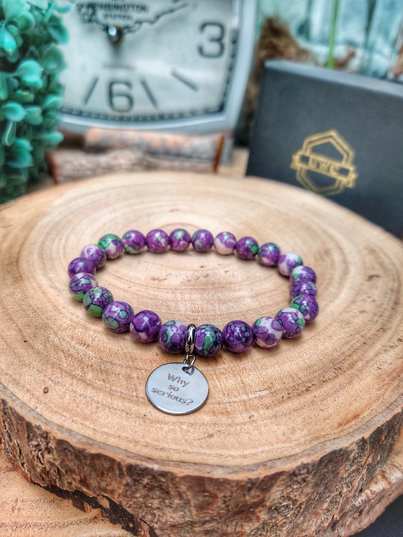 May include: A bracelet with purple and green speckled beads. A silver charm with the words "Why so serious?" is attached. The bracelet is presented on a wooden surface. The beads are approximately 0.8 cm in diameter.