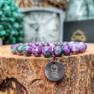 May include: A bracelet featuring purple and green beads, with a silver charm depicting a face. The bracelet is presented on a wooden surface. The background is blurred, with hints of green and white.