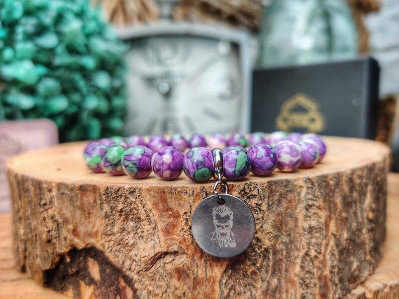 May include: A bracelet featuring purple and green beads, with a silver charm depicting a face. The bracelet is presented on a wooden surface. The background is blurred, with hints of green and white.