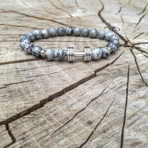 May include: A gray and white beaded bracelet with a silver dumbbell charm. The bracelet is on a wooden surface.