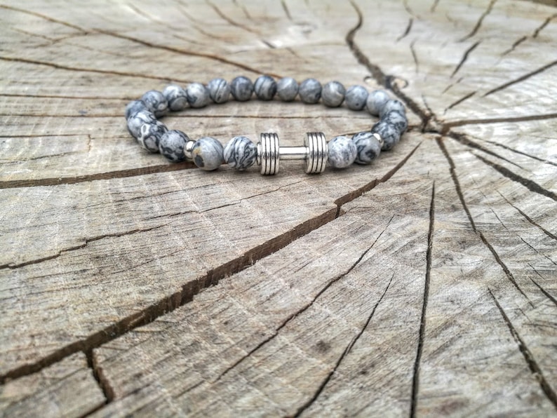 May include: A gray and white beaded bracelet with a silver dumbbell charm. The bracelet is on a wooden surface.