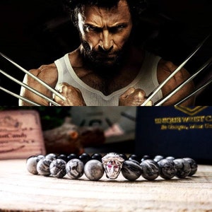 Op de afbeelding: Een armband van zwart en grijs marmer met een zilveren leeuwenkopbedel. De bedel heeft een rode steen als oog. De armband ligt op een houten oppervlak.