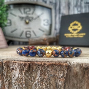 May include: A blue and brown beaded bracelet with a gold bead in the center. The bracelet is on a wooden surface.