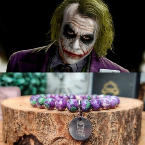 May include: A bracelet with purple and green beads and a silver charm with a Joker design. The bracelet is displayed on a wooden surface, with a portrait of the Joker in the background.