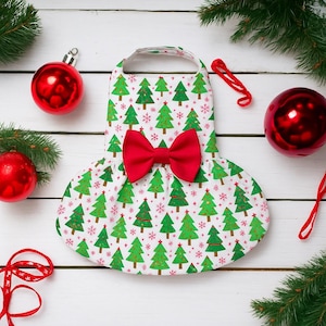 Puede incluir: Un vestido blanco para mascotas con un estampado de árbol de Navidad y un lazo rojo. El vestido presenta árboles de Navidad verdes y copos de nieve rojos. Adornos navideños rojos y ramas de hoja perenne en el fondo.