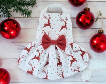 Puede incluir: Un vestido blanco con un lazo rojo y un estampado de renos. El vestido está decorado con pequeñas estrellas rosas y grises. El vestido está rodeado de adornos navideños rojos.