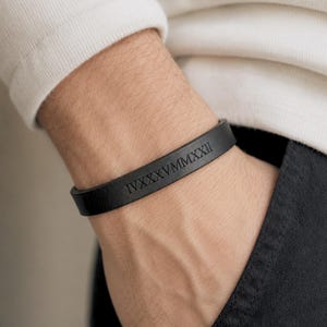 May include: A black leather bracelet featuring the Roman numerals IVXXXVMMXXII embossed in a clean, sans-serif font. The band has a smooth, matte finish and a simple, flat profile that wraps around the wrist with a secure, minimalist design.