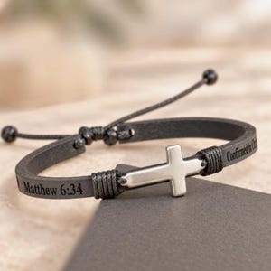 May include: A dark gray leather bracelet featuring a central silver-toned metal cross. The band is engraved with the text Matthew 6:34 and Confirmed in Christ. It includes adjustable black cord ties with small spherical beads at the ends.