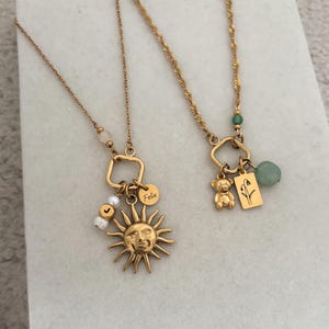 Può includere: Due collane color oro con ciondoli. Una presenta un sole con un volto, un disco con il nome "Felix" e perle bianche. L'altra ha un orsetto, un'etichetta floreale e una pietra verde.