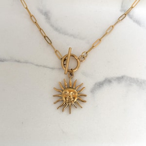 Può includere: Collana dorata con un ciondolo a forma di sole con un volto sorridente. La collana ha una chiusura a levetta e una catena delicata. Il ciondolo a forma di sole è dettagliato con raggi che si estendono verso l'esterno. Il gioiello è esposto su una superficie di marmo.