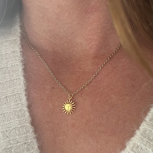 Könnte beinhalten: Eine goldene Kettenkette mit einem kleinen Sonnenanhänger. Die Sonne hat ein Gesicht und Strahlen.