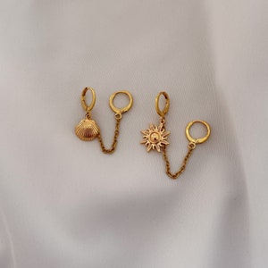 Peut inclure: Boucles d'oreilles créoles dorées avec une chaîne pendante reliant un charme coquillage à un charme soleil.