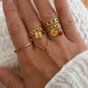 Könnte beinhalten: Eine Sammlung goldfarbener Ringe, darunter ein Ring mit Kettengliedern und einem rechteckigen Anhänger mit dem Buchstaben "D", ein Ring mit einem Anhänger und ein Ring mit einer kleinen roten Perle. Eine zarte Goldkette verbindet die Ringe.